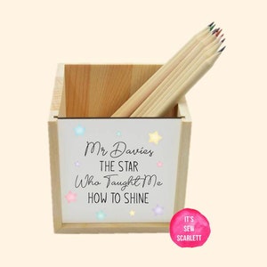 May include: A wooden pencil holder with a white front panel that says "Mr. Davies The Star Who Taught Me How To Shine" with colorful stars. The holder is filled with wooden pencils.