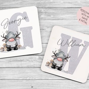 May include: Two white ceramic coasters with a gray glitter background. Each coaster features a gray and white reindeer gnome holding a gift. The first coaster has the letter G and the name Georgie. The second coaster has the letter W and the name William.