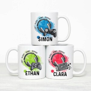 May include: Three white ceramic mugs with personalized names "Ethan", "Simon", and "Clara". Each mug features a watercolor-style design of a video game controller, headphones, and the text "Eat-Sleep-Game-Repeat".