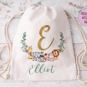 May include: A white drawstring bag with a gold letter E and the name Elliot printed in green. The bag features a wreath of eucalyptus leaves and flowers with a giraffe, elephant, zebra, and lion.