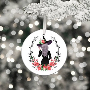 May include: A white round ceramic ornament with a black silhouette of a witch wearing a black hat and a floral wreath around her. The flowers are pink and white.