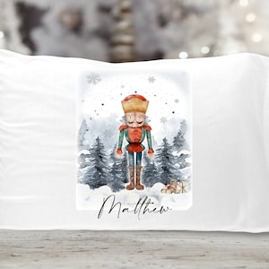 May include: White pillowcase with a watercolor illustration of a nutcracker standing in a snowy forest. The nutcracker is wearing a red and green outfit and a gold crown. The text "Matthew" is printed below the illustration.