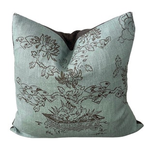 May include: A light blue throw pillow with a brown floral design. The pillow features a stylized tree with flowers and leaves, and a decorative pot at the base. The design is reminiscent of traditional Asian art.