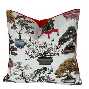 Puede incluir: Una almohada blanca con un borde rojo presenta un patrón repetido de árboles bonsái en varias macetas y un árbol de magnolia en flor. Los árboles están representados en un estilo japonés tradicional.