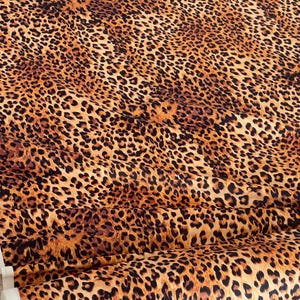 May include: A close-up of a fabric with a leopard print pattern. The fabric features a tan background with dark brown spots of varying sizes. The fabric is folded, showing the texture and pattern in detail.