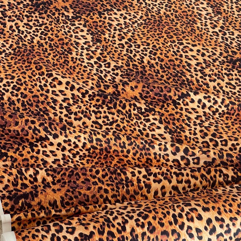 May include: A close-up of a fabric with a leopard print pattern. The fabric features a tan background with dark brown spots of varying sizes. The fabric is folded, showing the texture and pattern in detail.