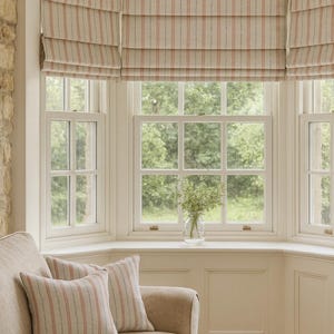 Neutral living room with bay window featuring hand-made Roman blinds in soft beige and pink striped linen-look fabric, styled with matching cushions on a beige sofa and views of lush green garden outside.