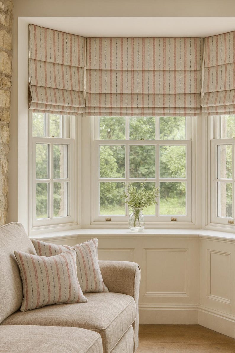 Neutral living room with bay window featuring hand-made Roman blinds in soft beige and pink striped linen-look fabric, styled with matching cushions on a beige sofa and views of lush green garden outside.