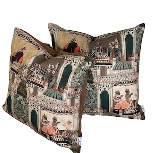 Puede incluir: Dos almohadas decorativas con fondo marr&oacute;n y estampado colorido de inspiraci&oacute;n india con personas, edificios y plantas.