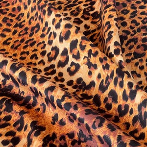 A close-up of a leopard print fabric. The fabric has a tan base with black and brown spots. The folds in the fabric create shadows, highlighting the texture and the pattern's details.