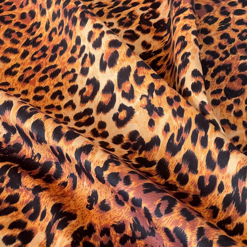 A close-up of a leopard print fabric. The fabric has a tan base with black and brown spots. The folds in the fabric create shadows, highlighting the texture and the pattern's details.