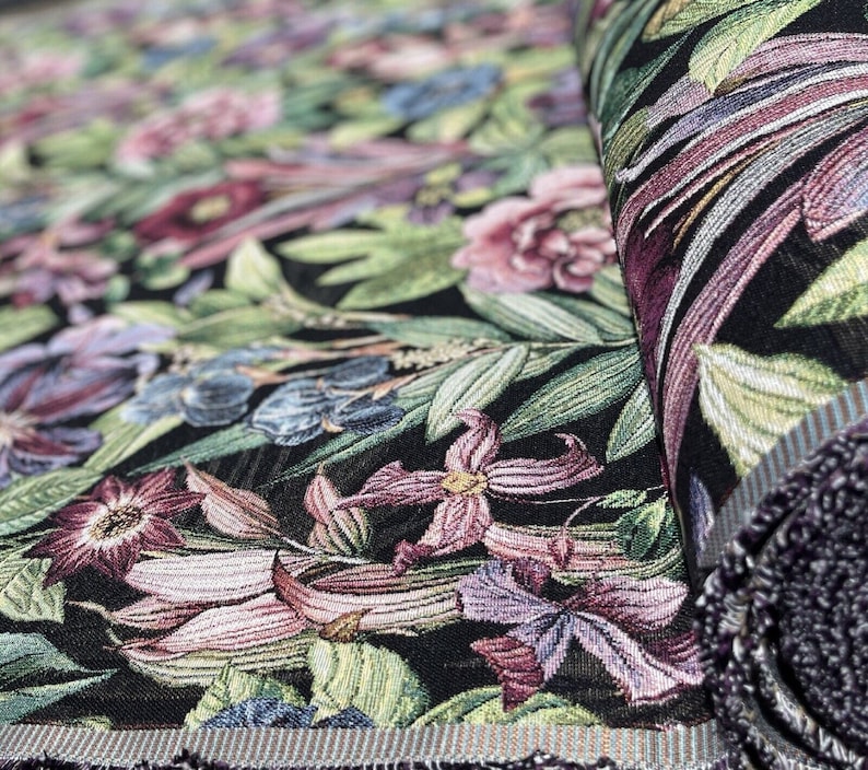 May include: Close-up of a black fabric with a floral tapestry design. The fabric features a variety of flowers in shades of pink, purple, and blue, as well as green leaves. The fabric is woven with a textured pattern.