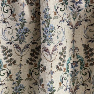 May include: Close-up of a beige fabric with a repeating floral pattern. The design features blue, green, and brown floral and leaf motifs. The fabric has a textured appearance, with visible woven threads.