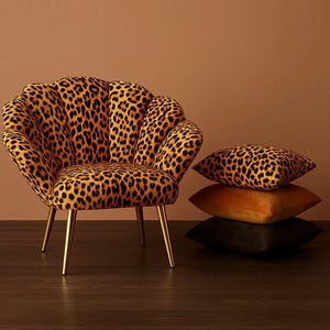 Luxurious velvet accent chair upholstered in a bold leopard-print fabric, featuring a scalloped shell-shaped backrest and slim gold metal legs. The chair sits on a dark wood floor against a warm tan backdrop. Beside it, three stacked decorative pillows—including a matching leopard-print velvet cushion—complement the rich, glamorous aesthetic.