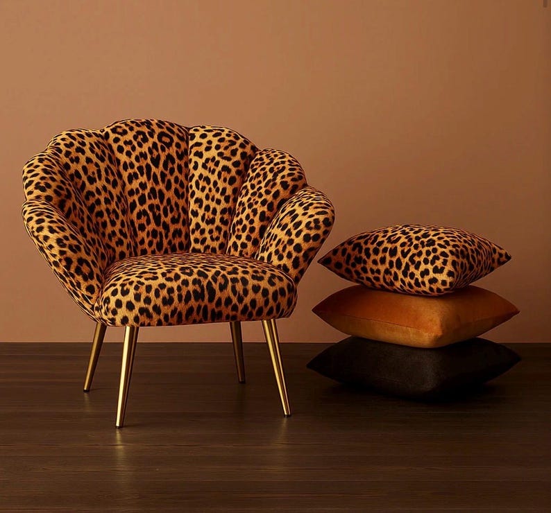 Luxurious velvet accent chair upholstered in a bold leopard-print fabric, featuring a scalloped shell-shaped backrest and slim gold metal legs. The chair sits on a dark wood floor against a warm tan backdrop. Beside it, three stacked decorative pillows—including a matching leopard-print velvet cushion—complement the rich, glamorous aesthetic.