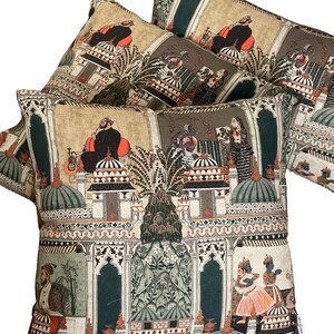 Puede incluir: Tres almohadas decorativas con un fondo beige y un patr&oacute;n repetido de ilustraciones verdes, naranjas y negras de personas y edificios. Las ilustraciones recuerdan al arte tradicional indio.