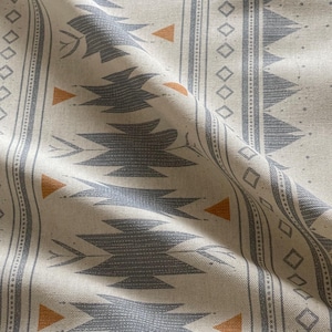 May include: Close-up of a beige fabric with a repeating pattern. The design features grey geometric shapes, orange triangles, and vertical stripes with diamond and dot details. The fabric has a textured appearance.