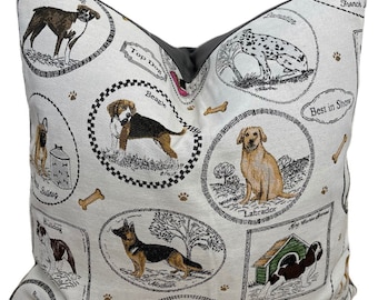 Peut inclure: Un coussin blanc avec un motif brodé coloré représentant différentes races de chiens, dont un Boxer, un Beagle, un Labrador, un Bulldog et un Jack Russel. Le coussin a une bordure grise et le texte "Top Dog", "Best in Show", et les noms de chaque race de chien.