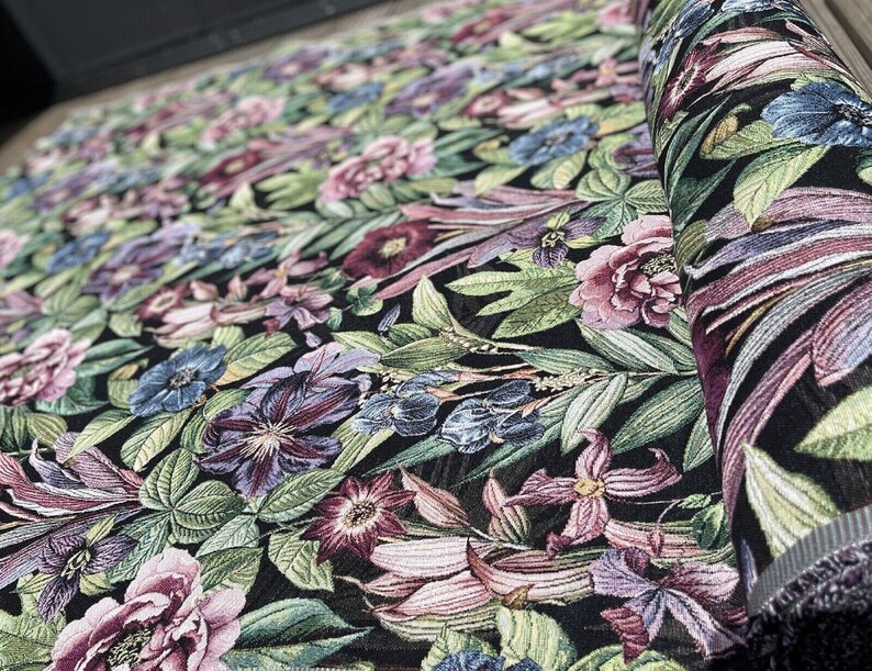 May include: Black fabric with a floral pattern featuring pink, purple, and blue flowers on a dark green background. The fabric is woven with a textured, raised design.