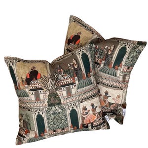 Puede incluir: Dos almohadas decorativas con fondo beige y un estampado colorido de inspiraci&oacute;n india con personas, edificios y plantas. El estampado es en tonos rojos, naranjas, verdes, azules y negros.