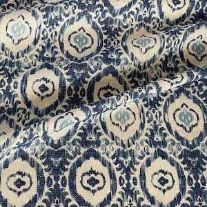 May include: A close-up of a fabric with a repeating pattern in shades of blue and cream. The design features stylized, oval-shaped motifs with a central, lighter blue element, creating a textured, ikat-inspired look. The fabric is folded, showing its drape.