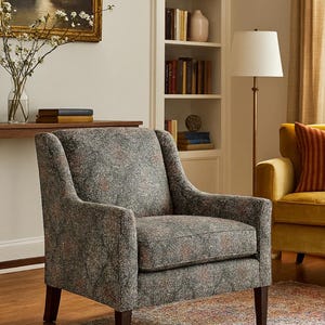 Puede incluir: Sillón tapizado con tela estampada en tonos grises, verdes y rojos. La silla tiene respaldo alto, reposabrazos y patas de madera oscura. La escena es un salón con una estantería y una lámpara de pie.