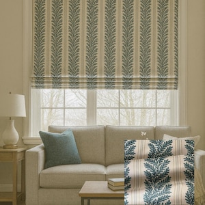 Peut inclure: Un store romain beige avec des rayures verticales et un motif répété de feuilles bleu-vert. Le store est installé au-dessus d'un canapé beige avec un coussin bleu. Une petite table en bois et une lampe sont à gauche.
