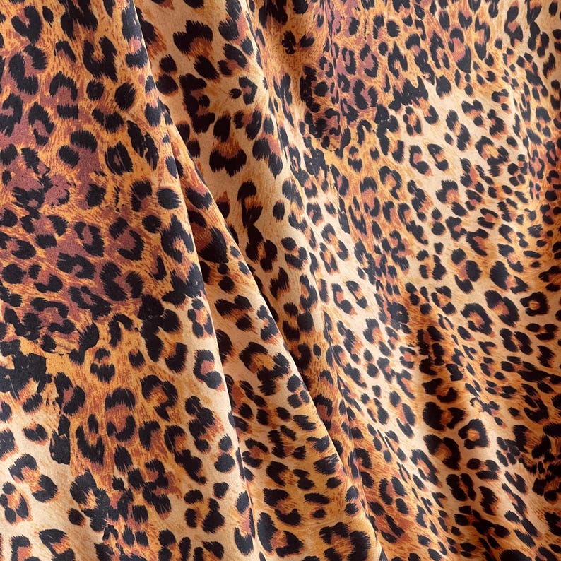 May include: Close-up of a fabric with a leopard print pattern. The fabric features a tan background with dark brown and black spots. The fabric is folded, creating shadows and depth, showcasing the texture and design.