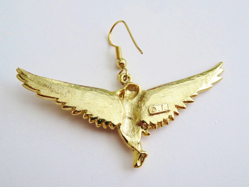 K&ouml;nnte beinhalten: Goldfarbene Ohrringe in Vogelform mit einem detaillierten Design. Die Ohrringe sind mit den Buchstaben "O.J.J." gekennzeichnet.