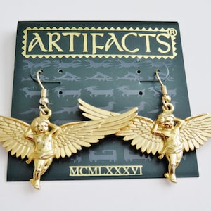 K&ouml;nnte beinhalten: Goldfarbene Engelsfl&uuml;gel-Ohrringe mit einer Cherubfigur an jedem Ohrring. Die Ohrringe befinden sich auf einer schwarzen Karte mit dem Wort "ARTIFACTS" in goldenen Buchstaben und dem Datum "MCMILXXXVI".
