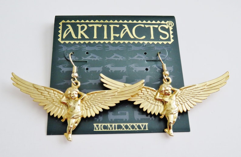 K&ouml;nnte beinhalten: Goldfarbene Engelsfl&uuml;gel-Ohrringe mit einer Cherubfigur an jedem Ohrring. Die Ohrringe befinden sich auf einer schwarzen Karte mit dem Wort "ARTIFACTS" in goldenen Buchstaben und dem Datum "MCMILXXXVI".