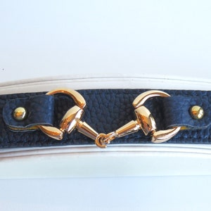 May include: A black leather bracelet with a gold-tone horse bit design. The bracelet has four gold-tone studs.