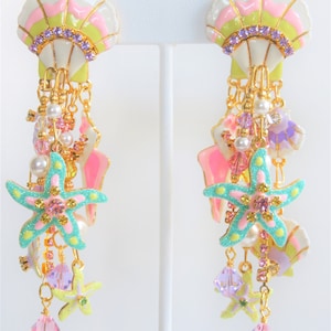 May include: A pair of seashell-themed earrings. The earrings feature a white, pink, and green seashell design with purple rhinestones. Dangling from the seashells are gold chains, pearls, and starfish in shades of turquoise, pink, and yellow.