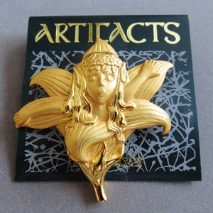 May include: A gold-colored brooch in the shape of a fairy within a flower. The brooch features the word "ARTIFACTS" in gold lettering on a black background. The fairy has long hair and is wearing a headpiece.