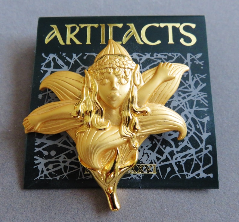 May include: A gold-colored brooch in the shape of a fairy within a flower. The brooch features the word "ARTIFACTS" in gold lettering on a black background. The fairy has long hair and is wearing a headpiece.