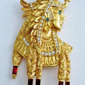 May include: A gold-tone brooch featuring a detailed profile of a woman with a crown and feathers. The brooch is adorned with clear and green rhinestones and has red dangling accents.