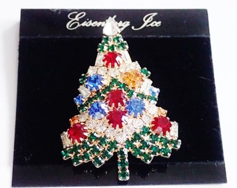 May include: A gold-tone Christmas tree brooch with green, red, blue, and clear rhinestones. The brooch is on a black velvet background and has the text "Eisenberg Ice" on the background.