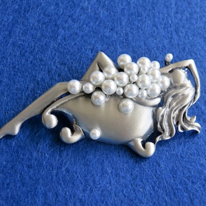 May include: A silver-toned metal brooch depicting a reclining figure in a bathtub, adorned with faux pearls. The figure has long flowing hair and is surrounded by a cluster of white pearls, creating a luxurious aesthetic.