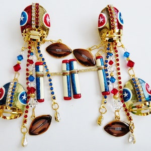 May include: A pair of gold football helmet earrings with red, white, and blue accents. Each earring features a football helmet with rhinestones, a football, and a set of football goal posts. The earrings are adorned with crystal and red and blue beads.