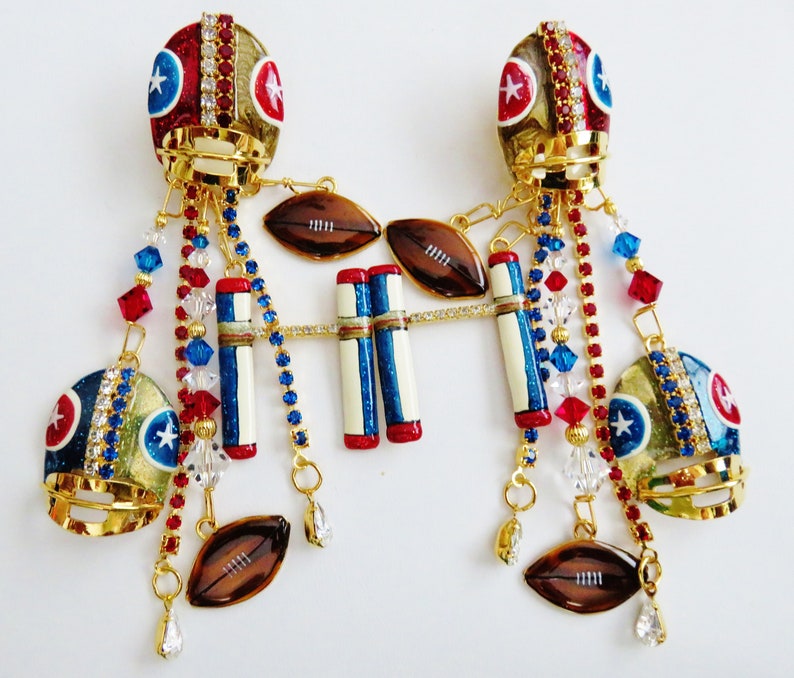 May include: A pair of gold football helmet earrings with red, white, and blue accents. Each earring features a football helmet with rhinestones, a football, and a set of football goal posts. The earrings are adorned with crystal and red and blue beads.