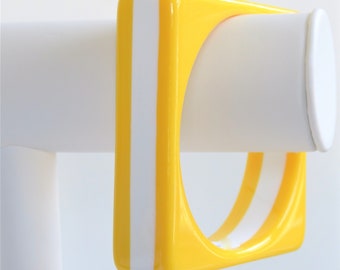 May include: A yellow and white plastic bangle bracelet with a square shape. The bracelet is on a white display stand.