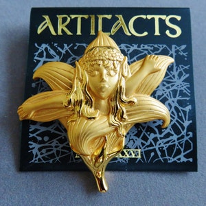May include: A gold-colored metal brooch in the shape of a fairy with a flower-like design. The brooch has the word "ARTIFACTS" in gold lettering on a black background.