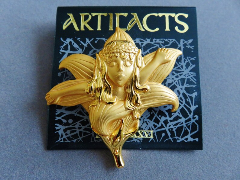 May include: A gold-colored metal brooch in the shape of a fairy with a flower-like design. The brooch has the word "ARTIFACTS" in gold lettering on a black background.