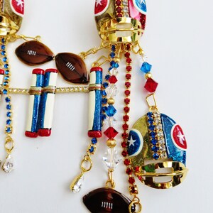 May include: Gold tone football helmet earrings with red, white, and blue details. The earrings feature footballs, goal posts, and crystal accents.