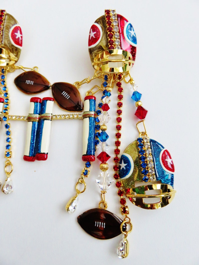May include: Gold tone football helmet earrings with red, white, and blue details. The earrings feature footballs, goal posts, and crystal accents.