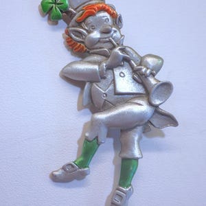 Puede incluir: Una figurita de peltre de color plateado de un duende tocando un instrumento musical. El duende lleva un sombrero verde, pantalones verdes y zapatos verdes. Tiene el pelo rojo y un trébol verde en el sombrero.