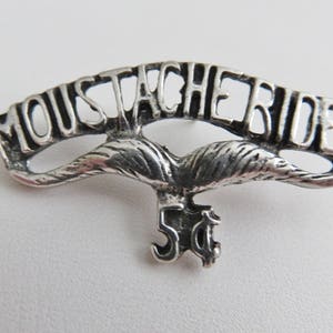 May include: Silver-toned pin with the words "Moustache Rides" in a curved banner design. A stylized mustache is below the banner, and the number "5¢" is below the mustache.
