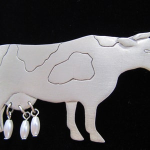 May include: A silver-toned cow brooch with a brushed finish. The cow has white patches and three dangling pearl-like charms. The brooch is a whimsical accessory, perfect for adding a touch of farm-inspired charm to any outfit.