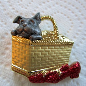 May include: A gold-colored metal pin depicting a dog sitting in a picnic basket with a red glitter shoe.