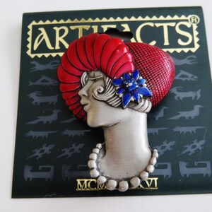 May include: A vintage-style brooch featuring a woman's profile with a red headdress and a blue flower accent. The brooch is silver-toned with a pearl necklace. The backing card reads "ARTIFACTS" and "MCM XVI".